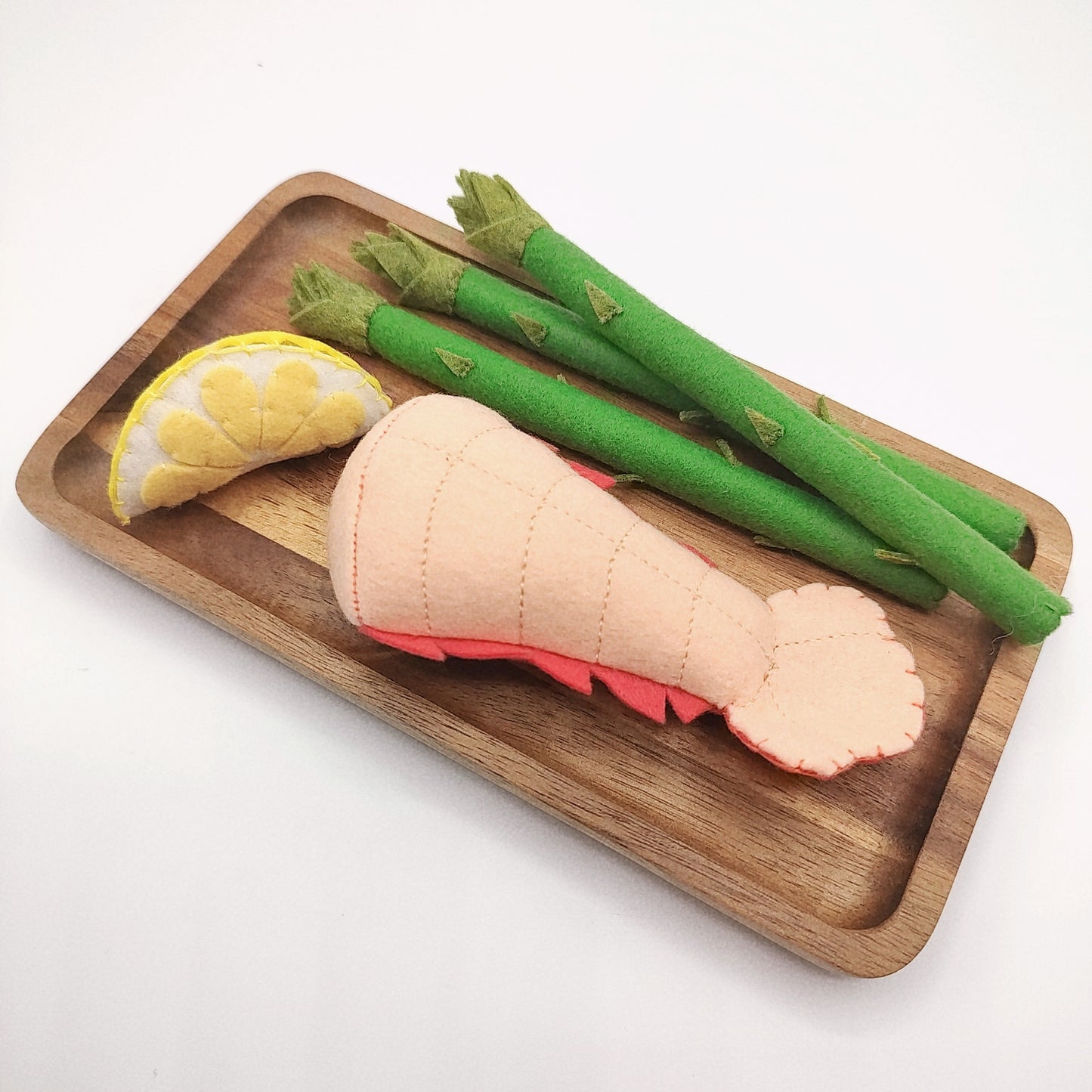 Wooden tray with felt food toys including salmon tail, green asparagus, and yellow lemon on a white background.