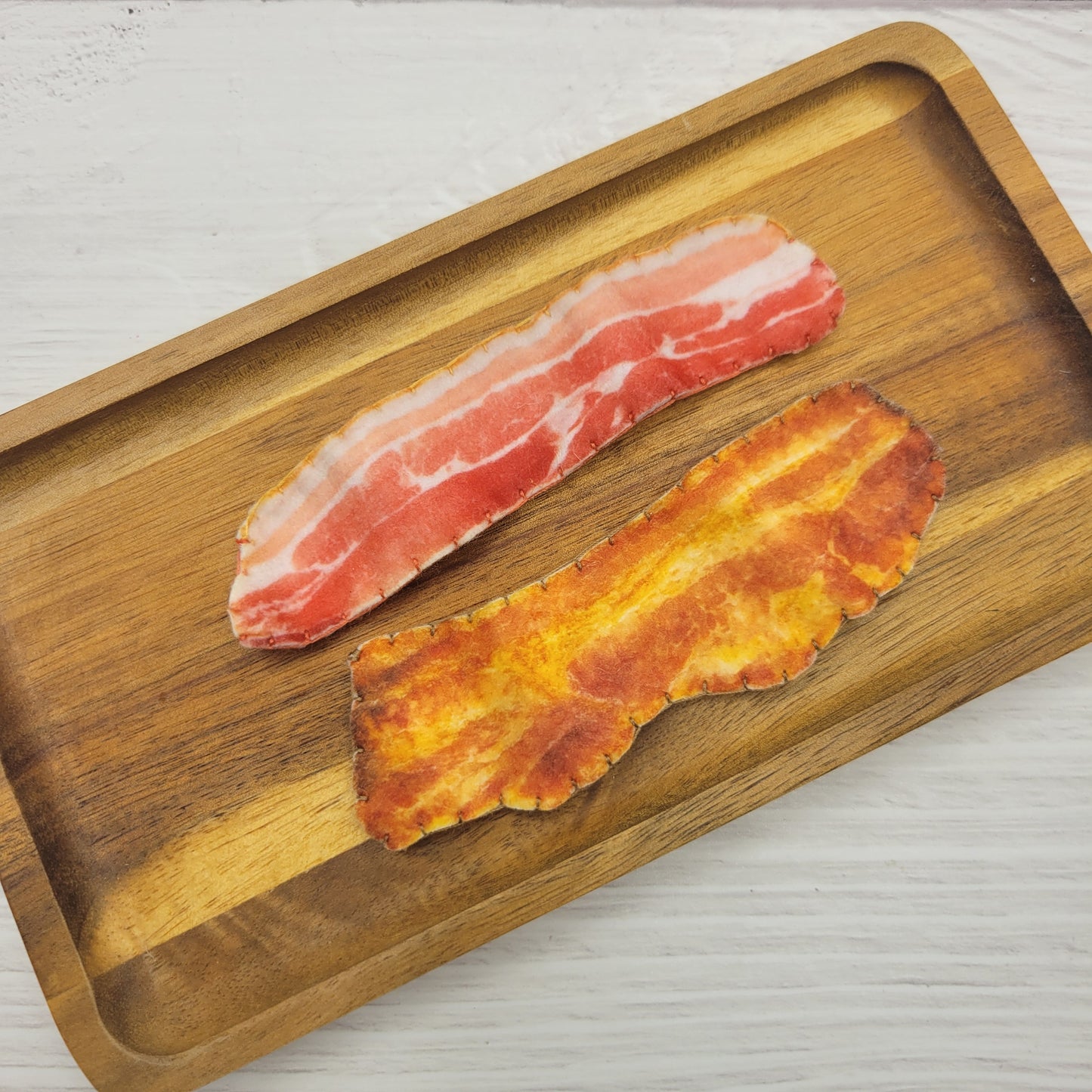 Two slices of pretend play plush bacon, one cooked and one raw, on a wooden tray.