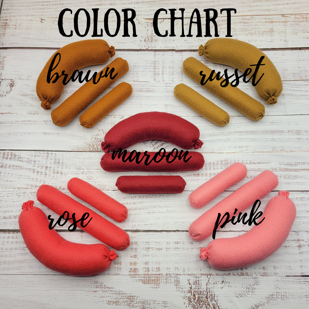 Color chart with plush pretend play toys sausages labeled 'brawn', 'russet', 'maroon', 'rose', and 'pink' on a wooden surface.