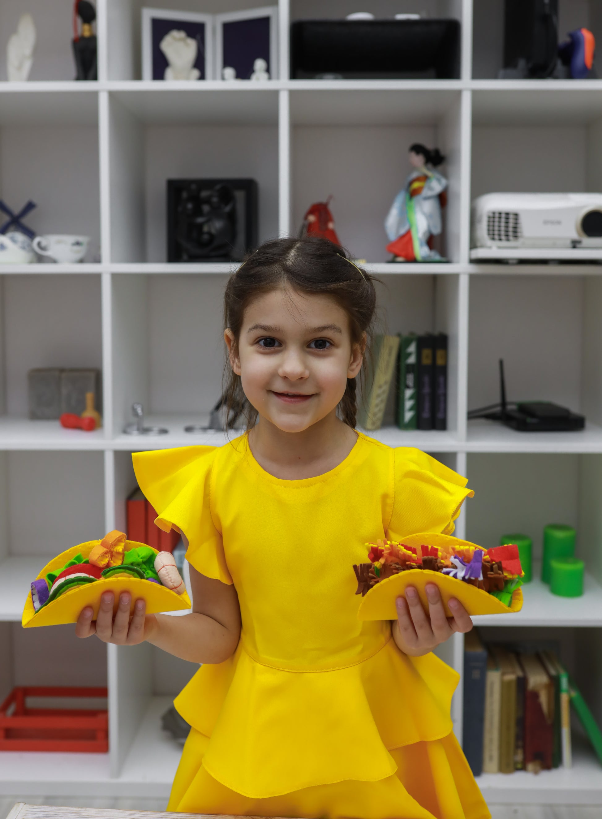 Young girl in a yellow dress holding colorful taco plush toys in front of a white shelf.